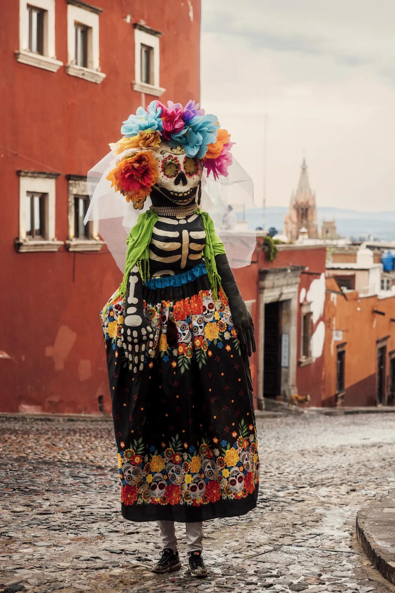 mojiganga figure dressed as a Catrina stands on a cobblestone street in San Miguel de Allende, wearing a colorful floral skirt, skeleton bodice, and a vibrant flower crown with a veil. The iconic Parroquia de San Miguel Arcángel is visible in the background.