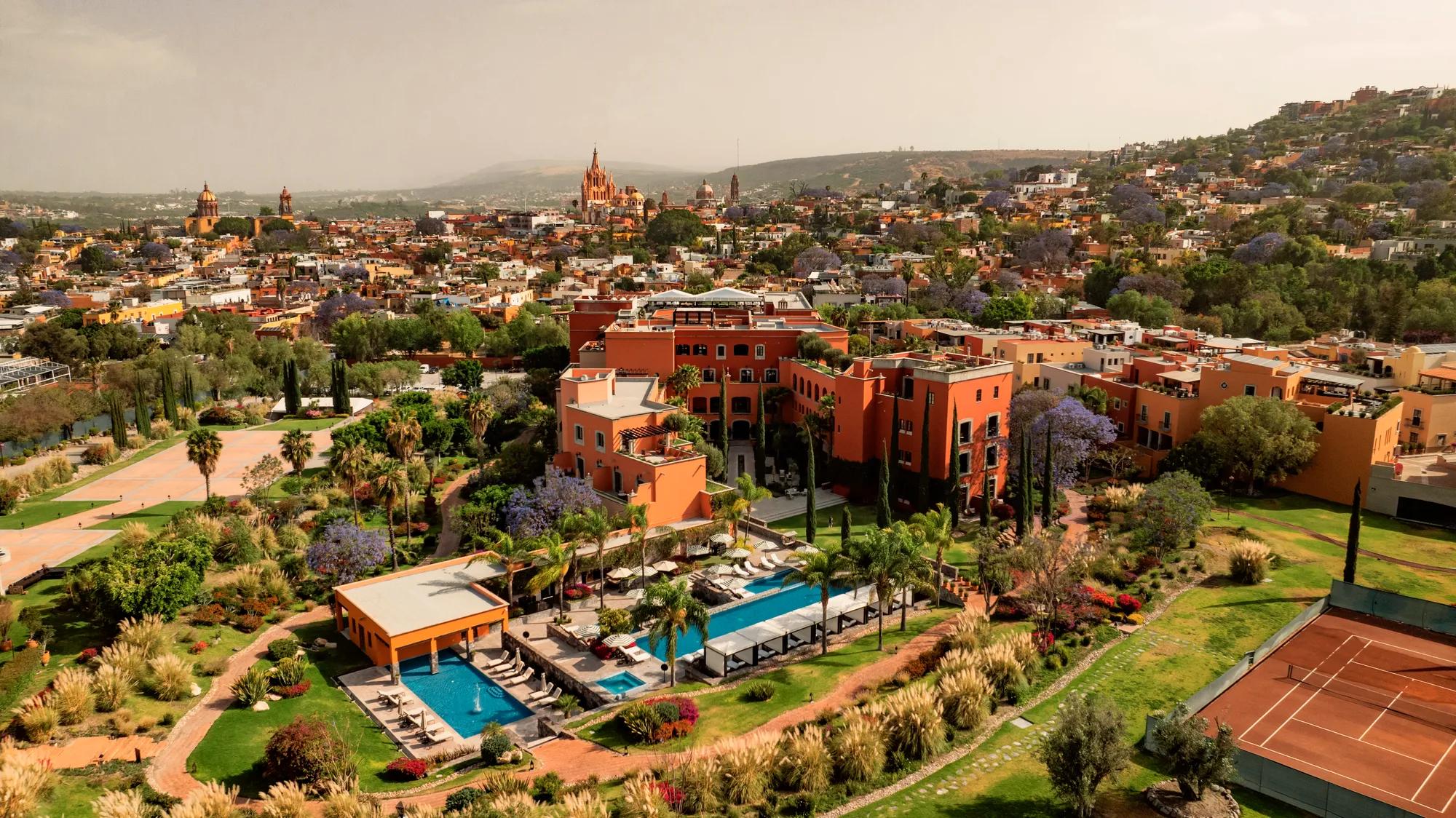 Aerial view of Rosewood San Miguel de Allende showcasing the hotel’s façade, pools, lush gardens, event spaces, and a panoramic backdrop of the iconic Parroquia de San Miguel Arcángel.