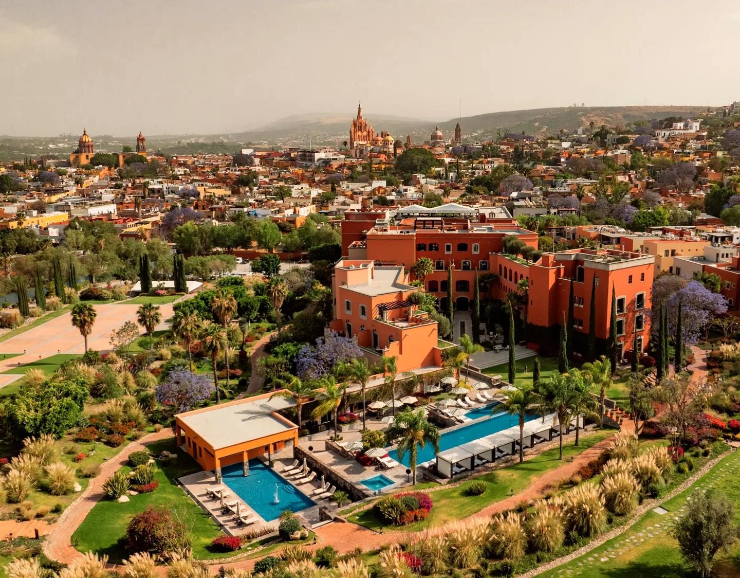 Aerial view of Rosewood San Miguel de Allende showcasing the hotel, pools with lounge chairs, gardens, event esplanade, tennis court, and residences, with the city and iconic Parroquia de San Miguel Arcángel in the background.