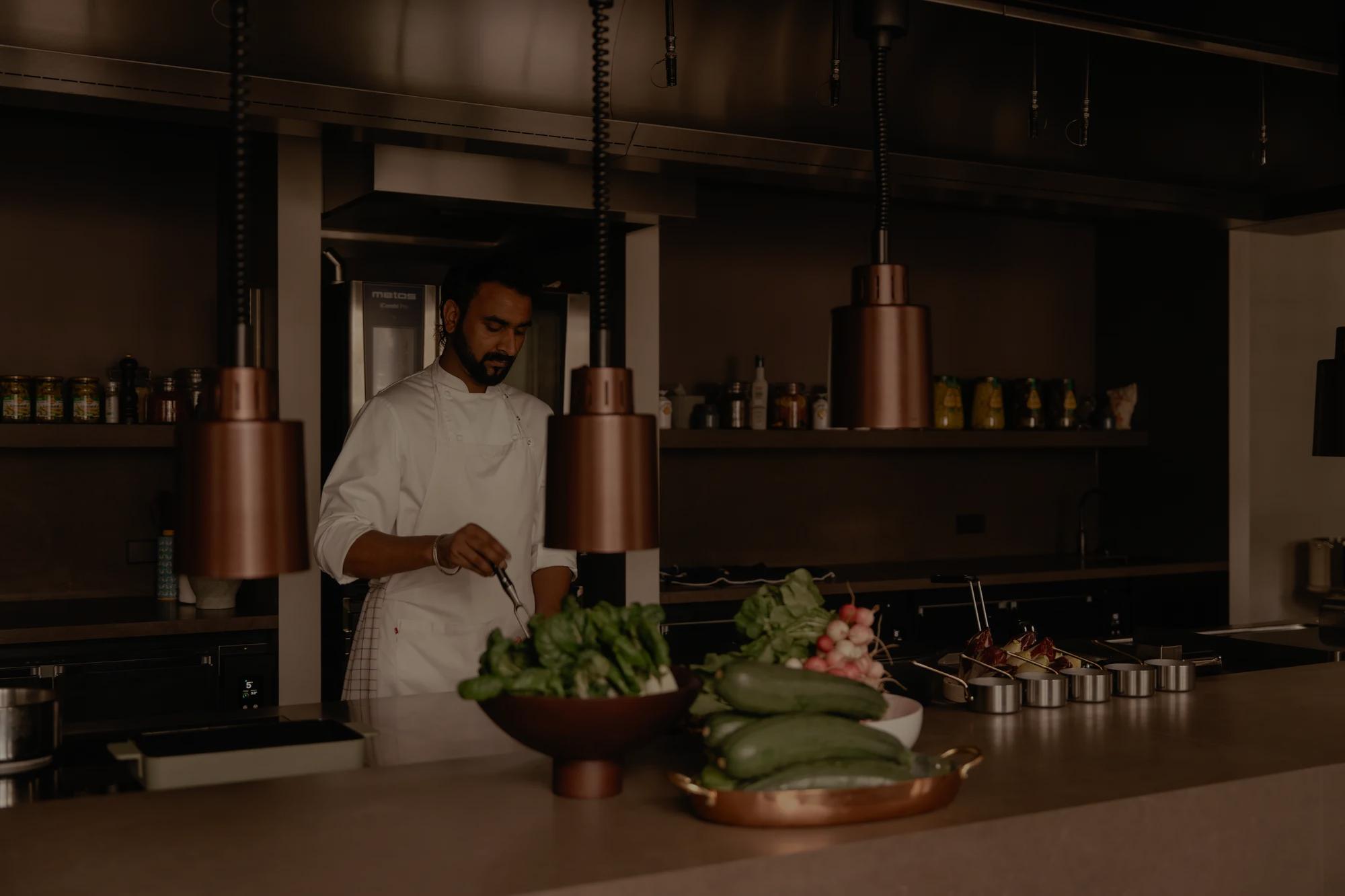 A chef prepares food in a modern kitchen with bronze pendant lamps and fresh vegetbales on the stone counter. 