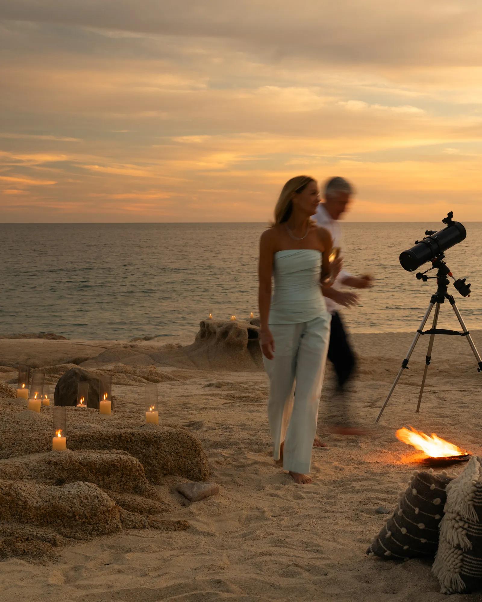 A couple walking along a sandy beach at sunset, with candles placed on the ground and a small fire nearby. A telescope stands on a tripod facing the ocean, and the sky is warm with orange and golden tones.