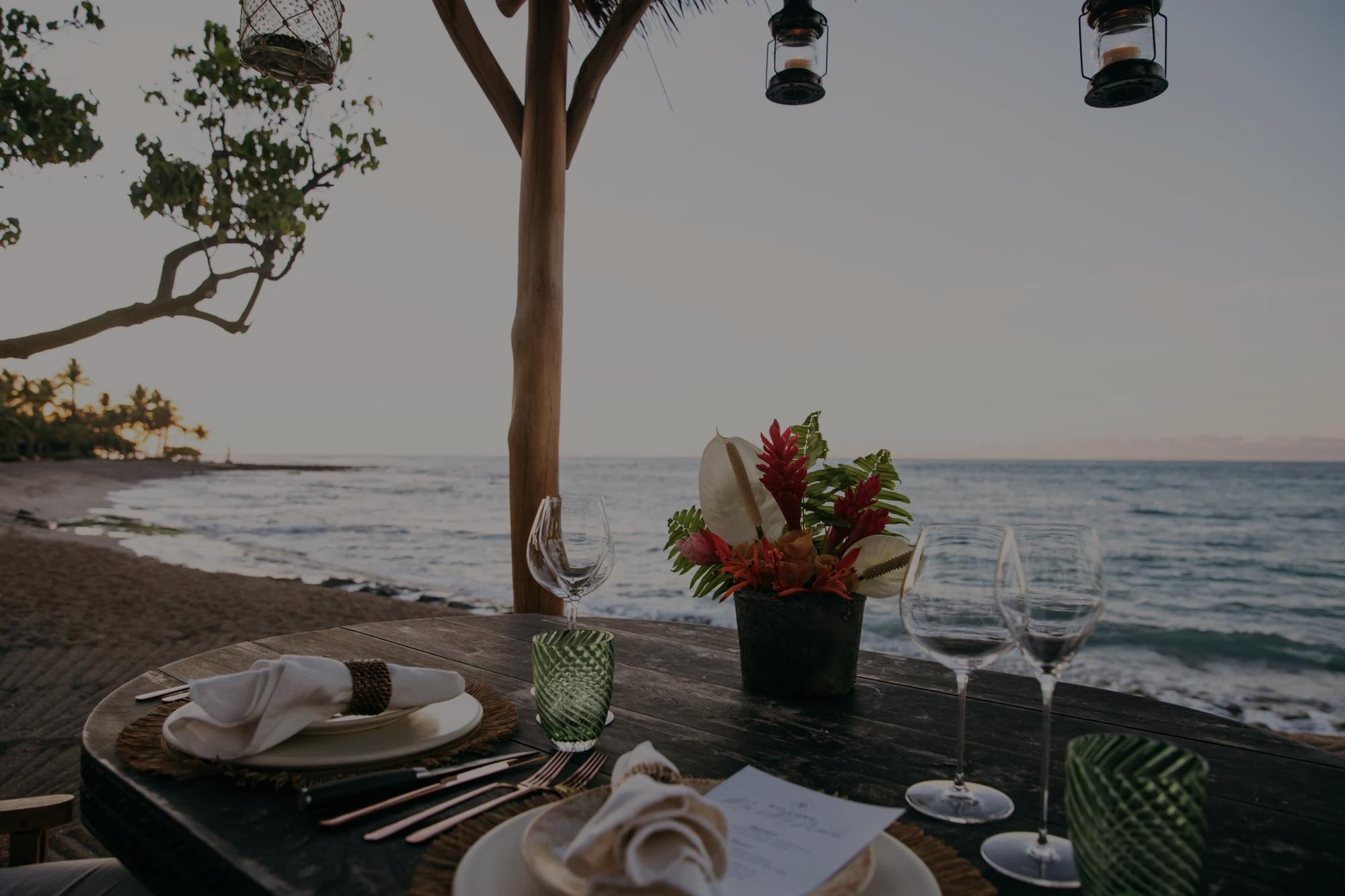 Wooden table on the beach at sunset with plates, silverware and glasses and florals