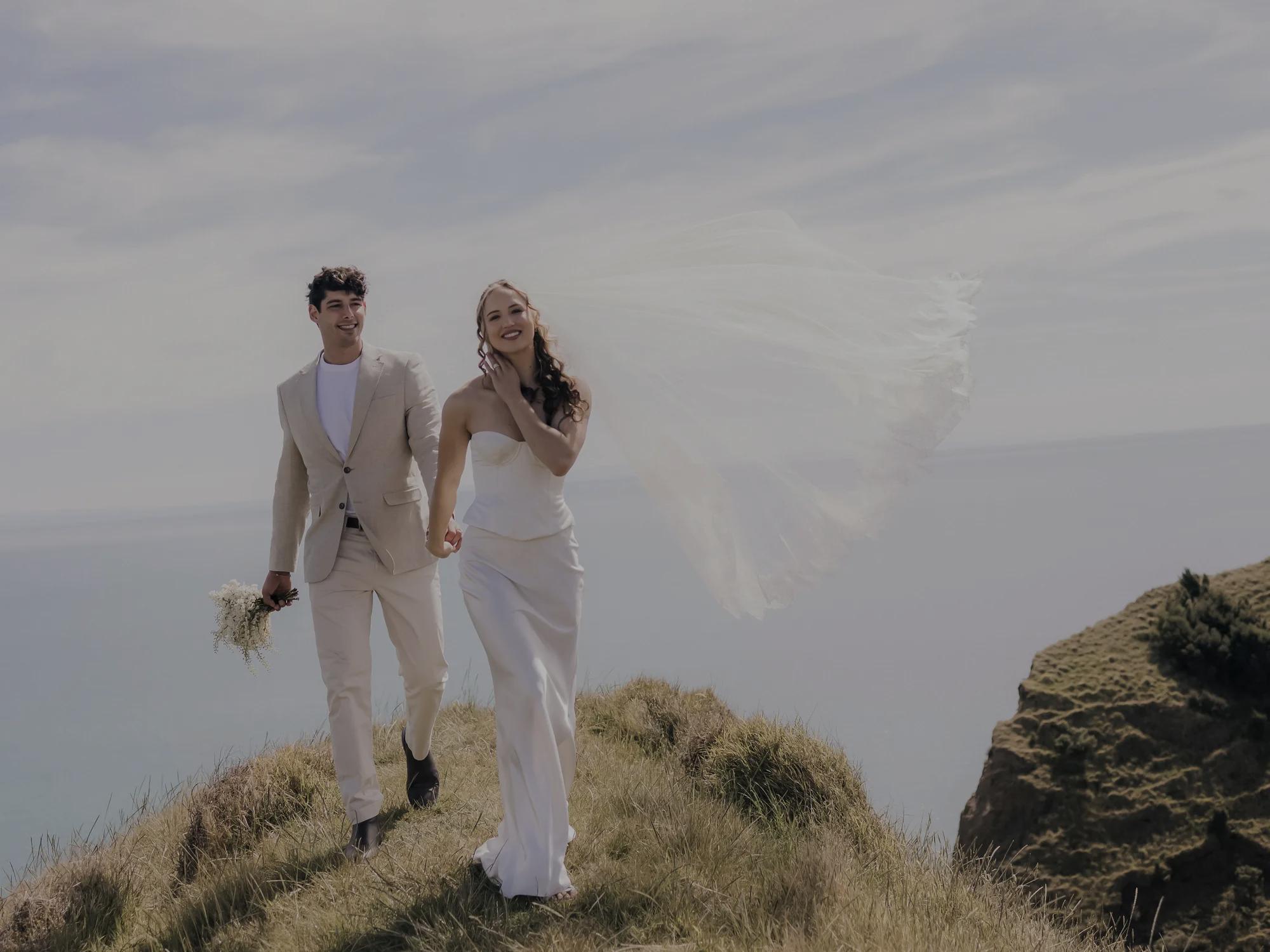 Romantic bride leading groom by the hand along a scenic ocean cliff, flowing wedding veil blowing in the wind.