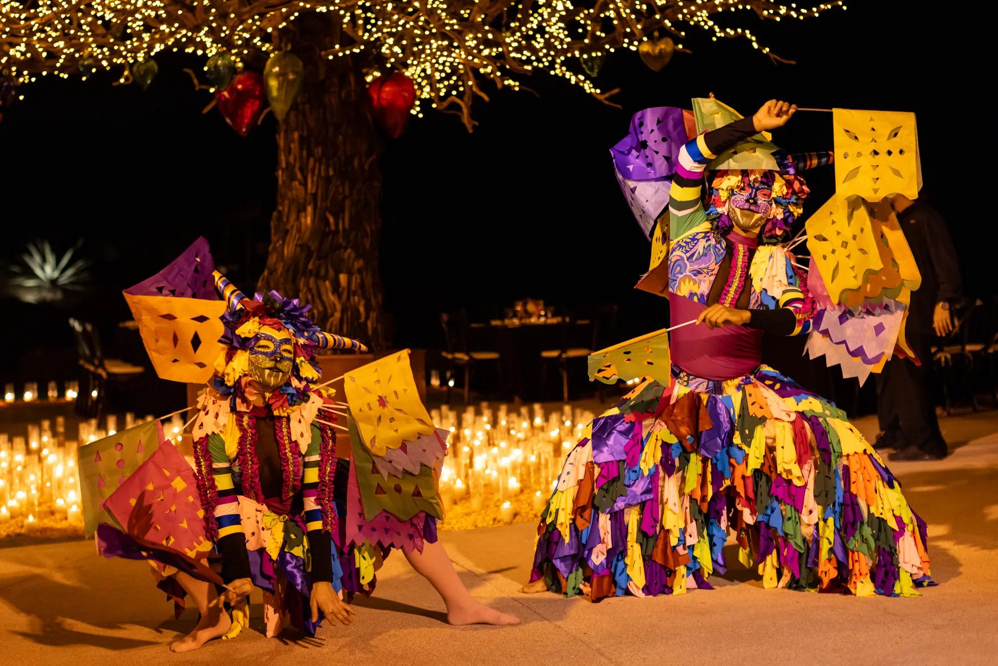 A woman in traditional folkloric dress dances gracefully, holding a decorative fan and lifting her lace skirt, under glowing golden lights. A man in a white outfit and hat stands smiling in the background, creating a festive Día de Muertos atmosphere.