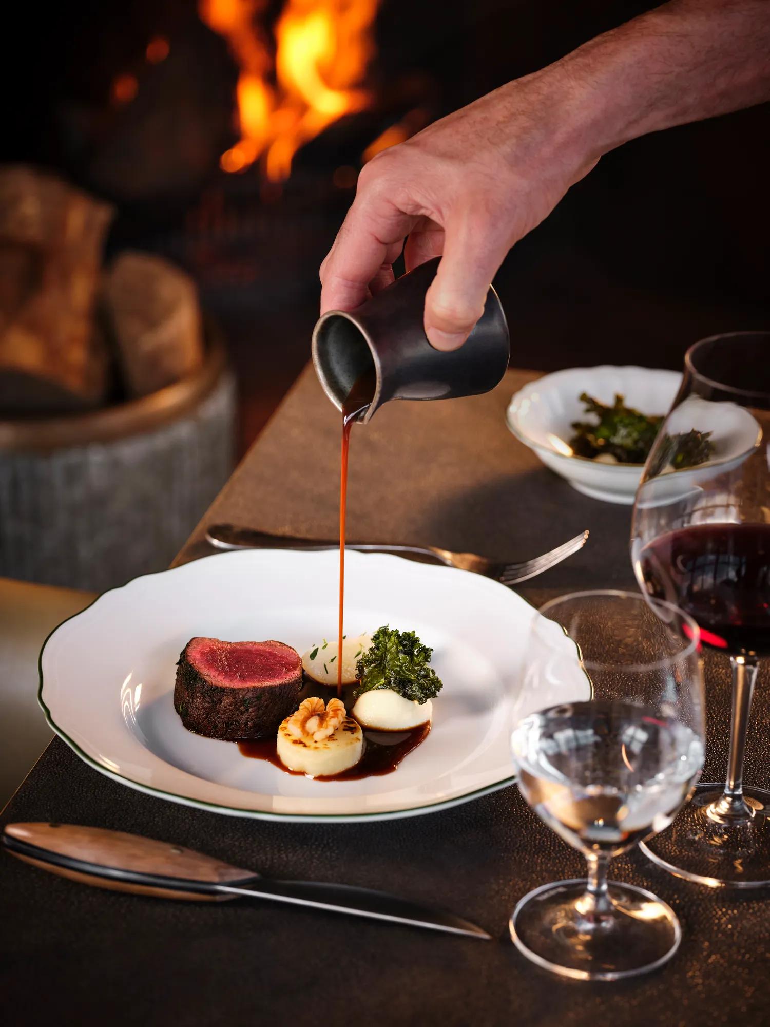 A hand pouring a dark sauce over a delicately presented venison and scallop dish in a restaurant setting