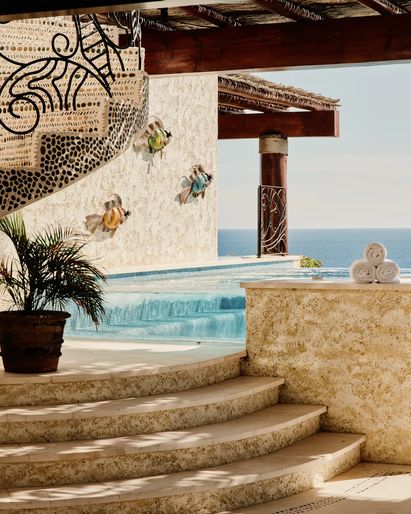 A sunlit terrace dining area with wooden table and chairs overlooking the ocean, framed by vibrant bougainvillea and a distant coastline.