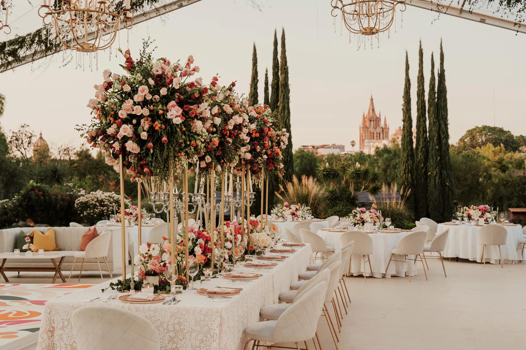 Wedding reception setup with pink tones in floral décor, linens, and seating, set outdoors with a transparent tent and the Parroquia in the background.