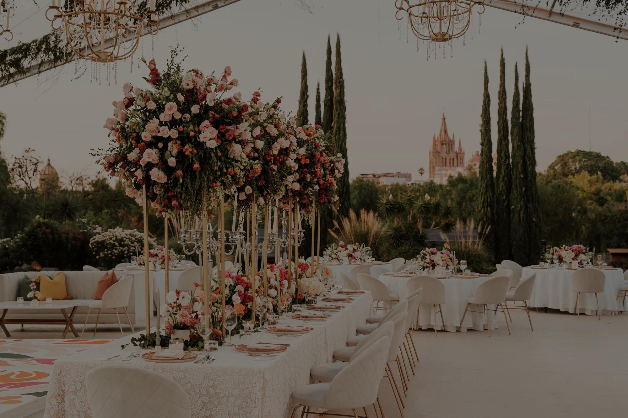 Wedding dinner setup with rectangular and round tables, white details, and pink floral arrangements, with the Parroquia de San Miguel Arcángel in the background.