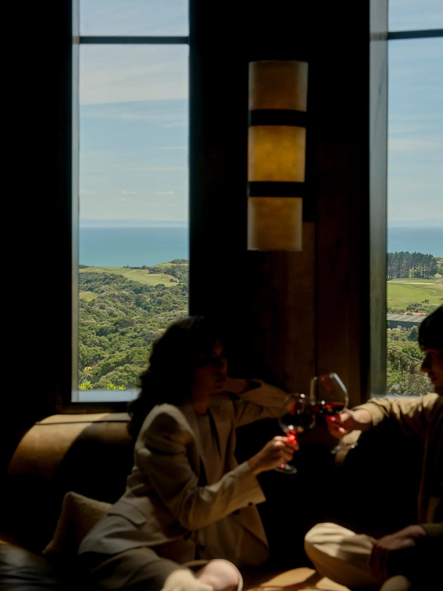A couple relax in a cozy room and cheers a glass of red wine, out the window are views of the property and ocean.