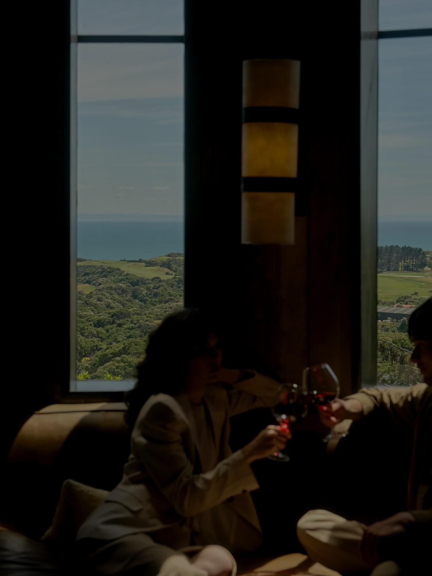 Couple cheers their glasses of red wine as they sit in a cosy snug with views of the ocean out the window.