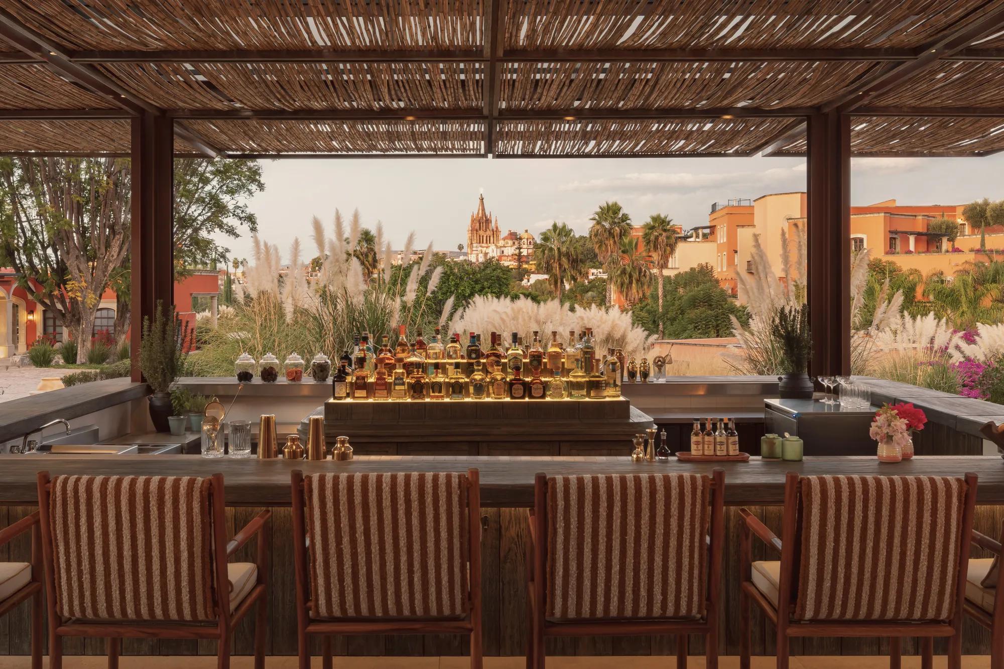 Outdoor rectangular wooden bar with four striped fabric-backed stools, a central island displaying bottles, set among plants with the iconic Parroquia de San Miguel Arcángel visible in the background.