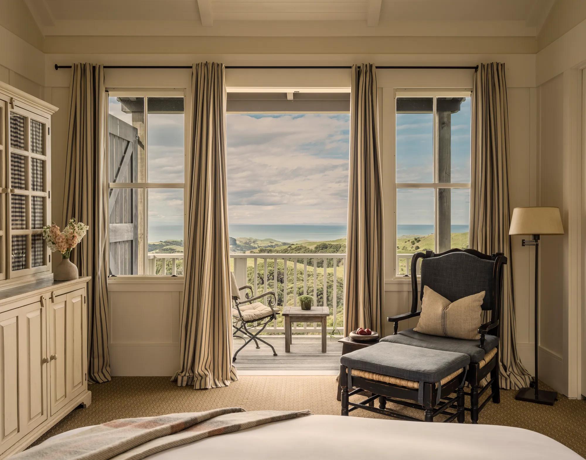 A hotel suite with neutral furnishings including a plush bed, chair and balcony with ocean views.