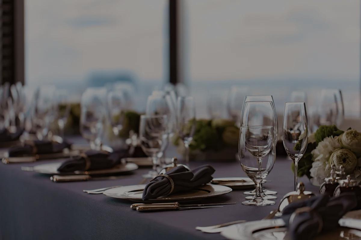 A formal dining table with a dark tablecloth, floral arrangements, and neatly arranged plates, silverware, and glasses, set against a backdrop of large windows.