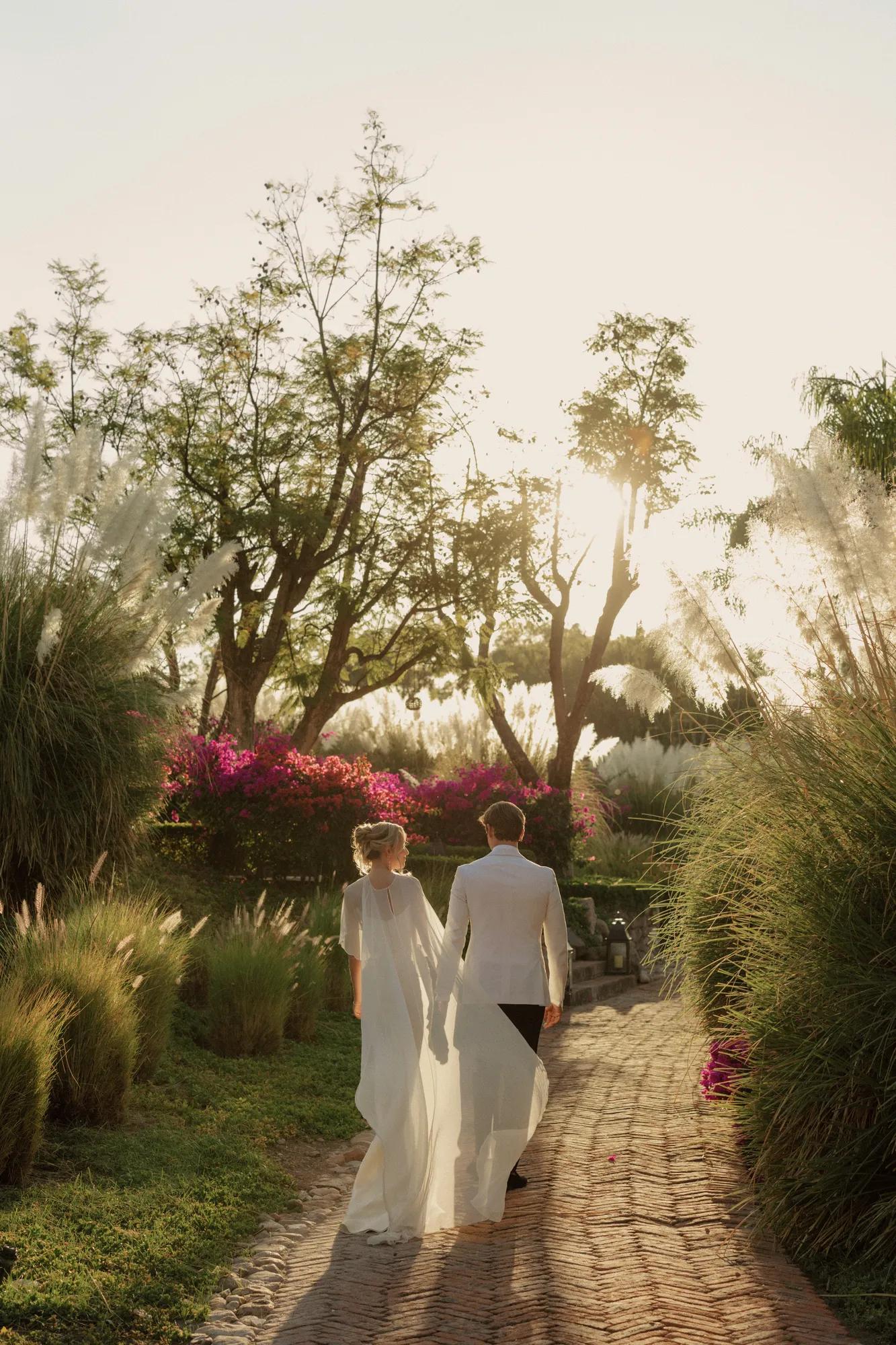 Bride and groom walking along a brick pathway surrounded by lush greenery, with the bride in a white gown and the groom in a suit.