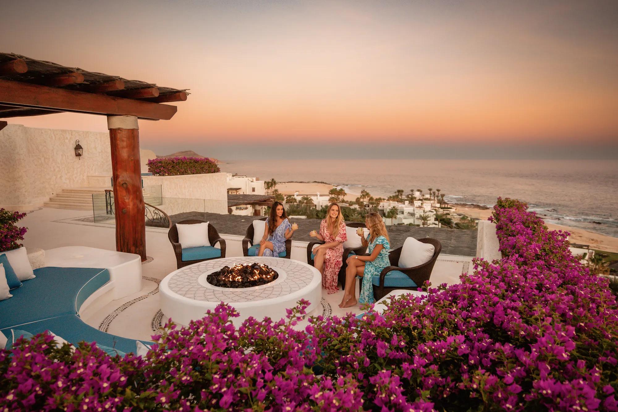 Three women sit around a circular fire pit on a terrace overlooking the ocean at sunset, surrounded by vibrant pink flowers and cushioned outdoor seating