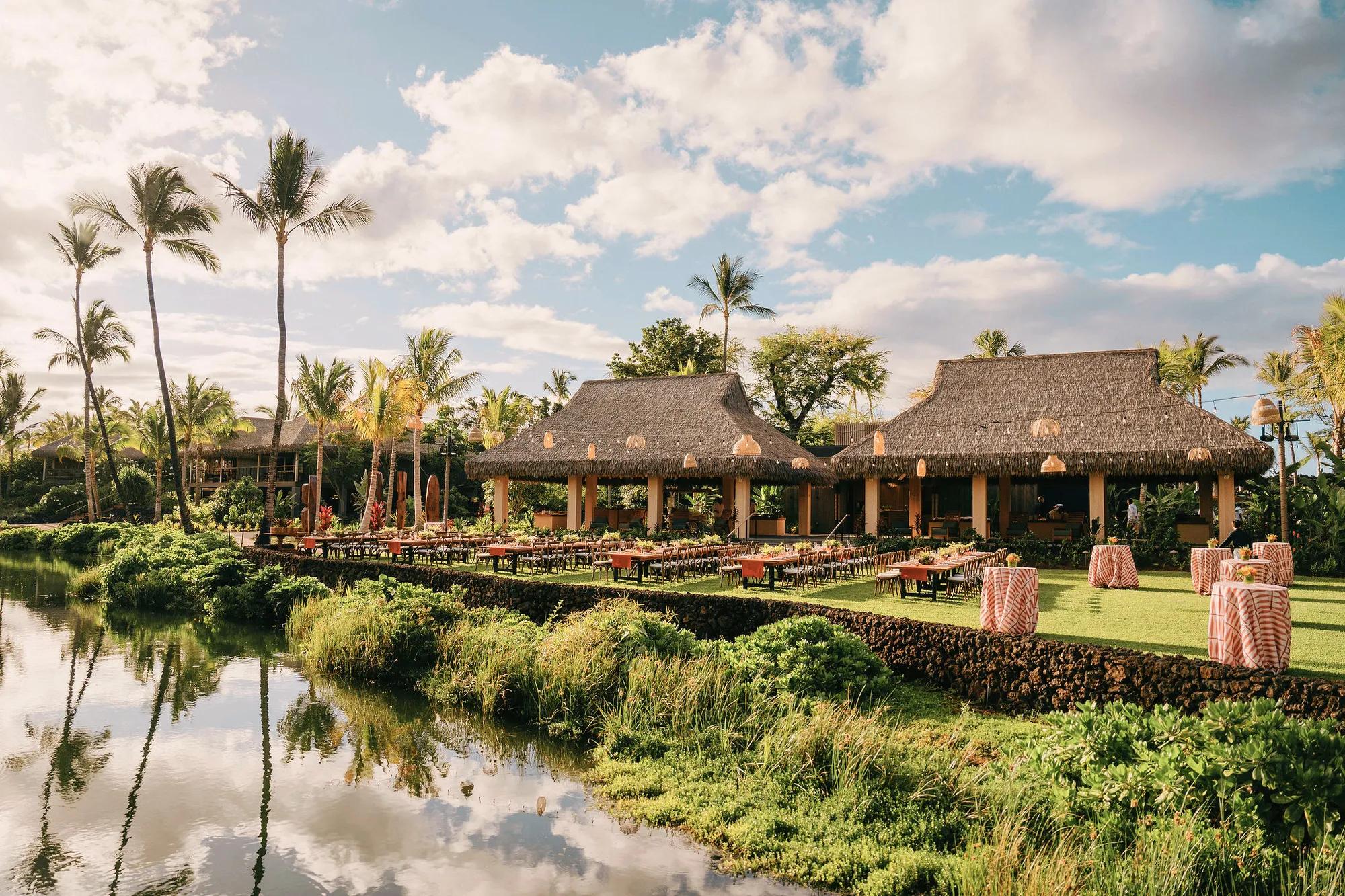 Expansive lawn with multiple tables covered in red and organge tablecloths with hanging string lights above and two thatched roof structures in the background
