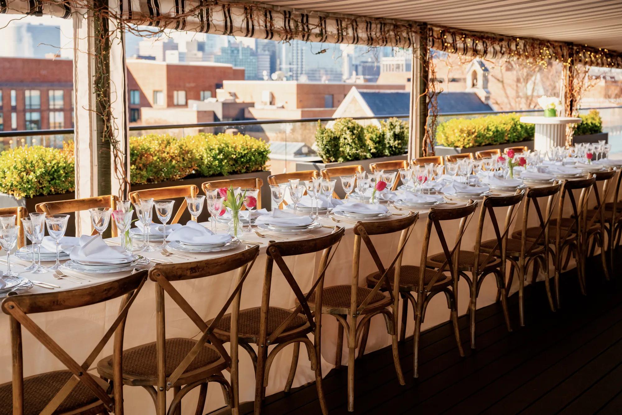 Long table set up for seated dinner on the rooftop with ivory linen and white hightops in the background.
