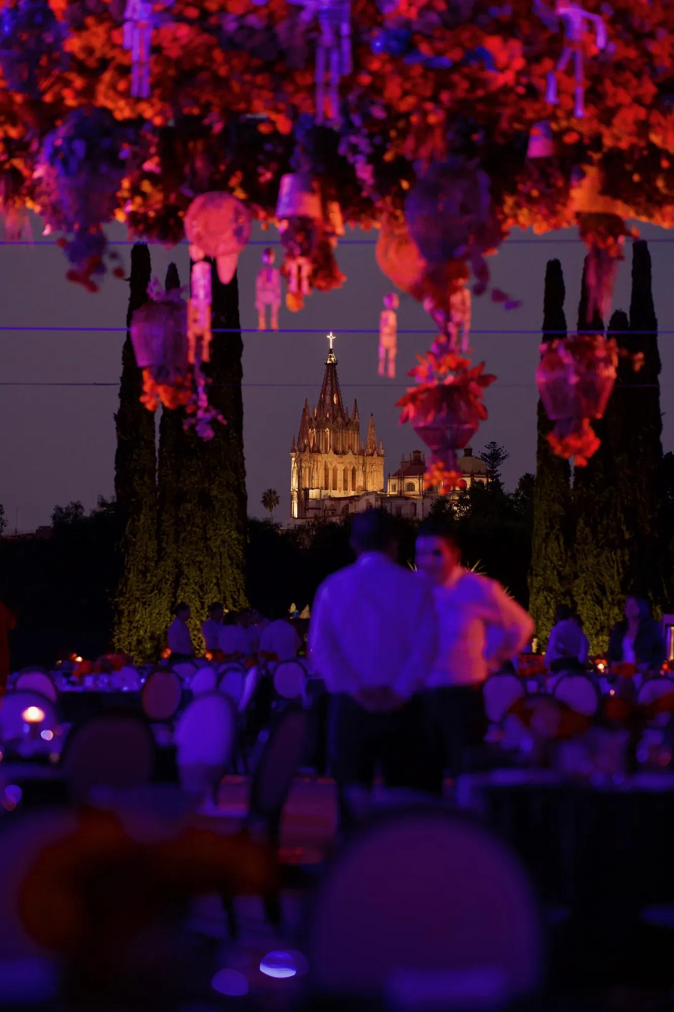 Event setup with a ceiling decorated with marigolds and skulls, multiple tables arranged on the ground floor, and the Parroquia de San Miguel Arcángel illuminated in the background among the trees at night.