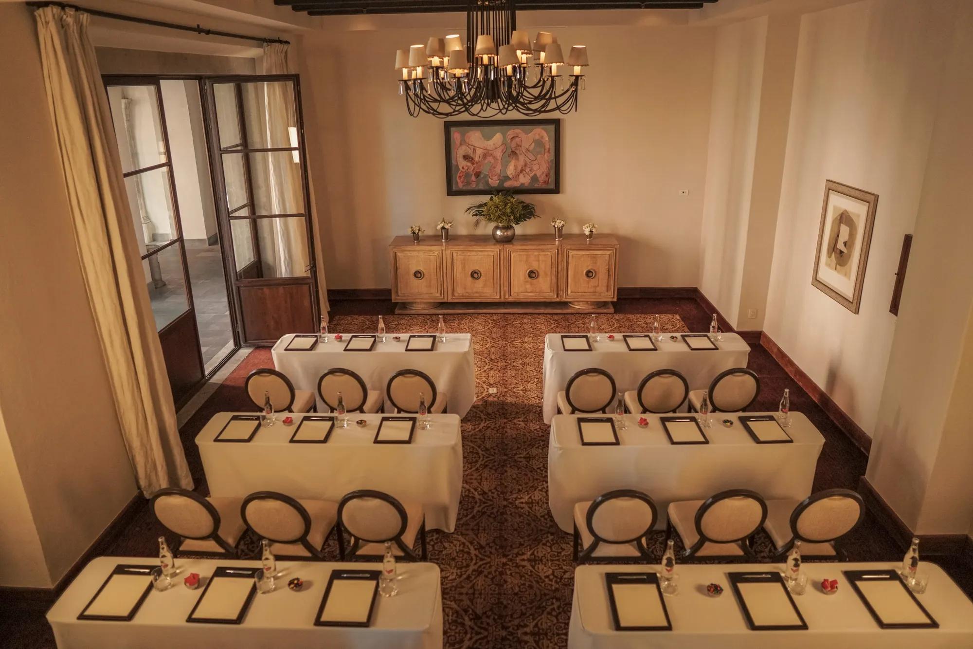 Meeting room with two rows of small round tables, each set with three chairs. A chandelier hangs from the ceiling, and a wooden-framed glass door stands open on the left side.