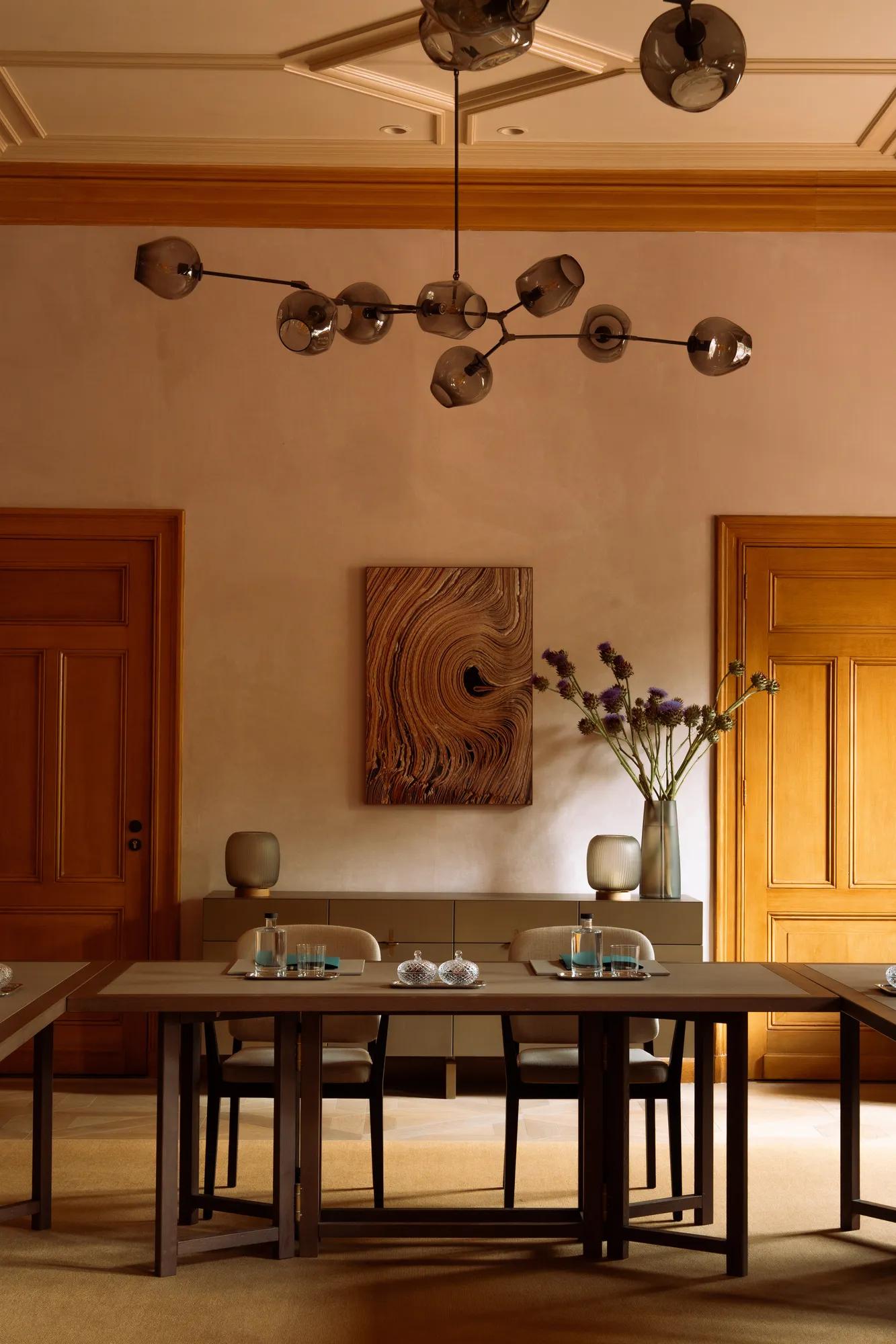 An elegant room with conference tables and chairs, with wooden doors and decorated with art, flowers and a chandelier. 