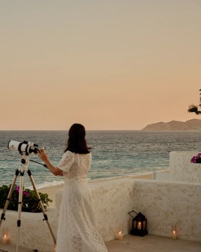 A seaside terrace at sunset with a telescope overlooking the ocean, surrounded by candles and floral arrangements, with palm trees and distant coastline under a warm orange sky.