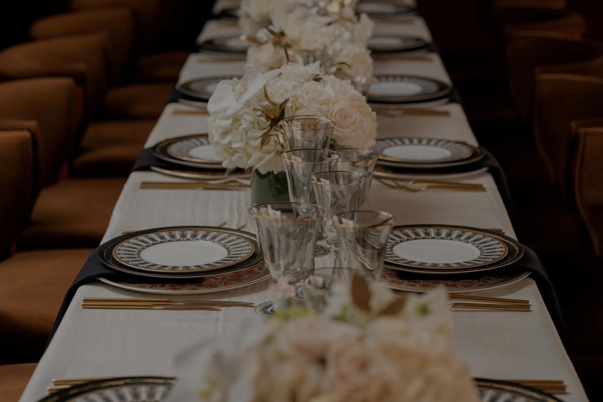 Elegant place setting with plates, glassware, and flatware on a long table with white linen and brown chairs. 