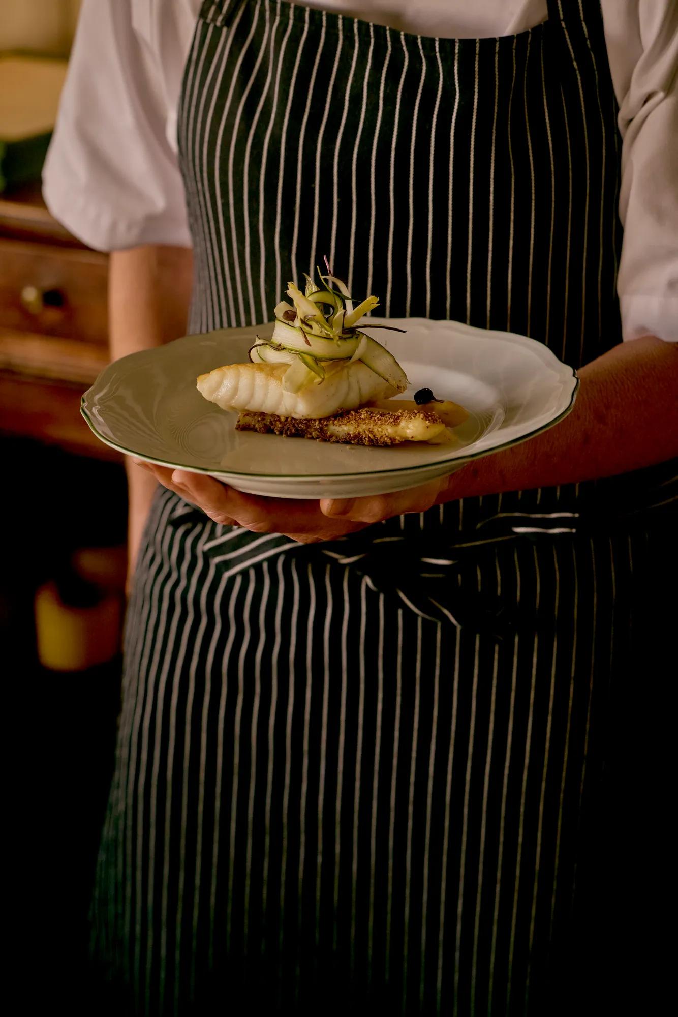 Chef in black and white pinstripe apron holding a plated gourmet fish and asparagus dish.